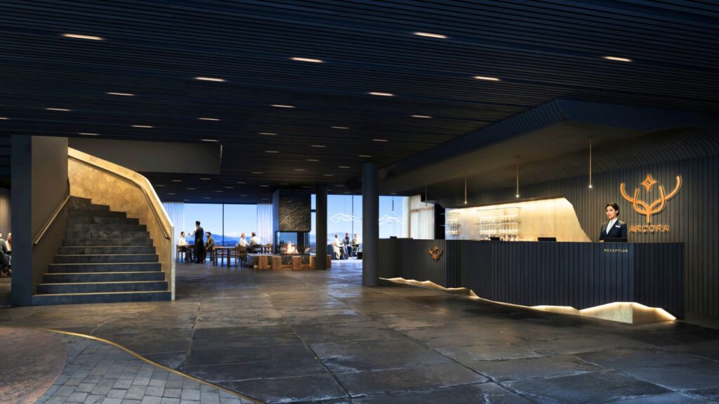 Modern hotel lobby with a black reception desk, logo, staircase on the left, and people seated near large windows in the background.