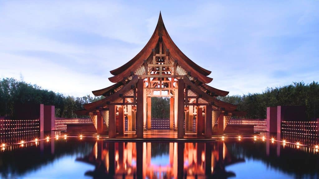 A wooden pavilion with a tiered roof stands over a reflective pool at Phulay Bay Krabi, surrounded by rows of small lights, with a clear sky in the background.