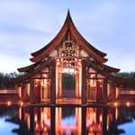 A wooden pavilion with a tiered roof stands over a reflective pool at Phulay Bay Krabi, surrounded by rows of small lights, with a clear sky in the background.