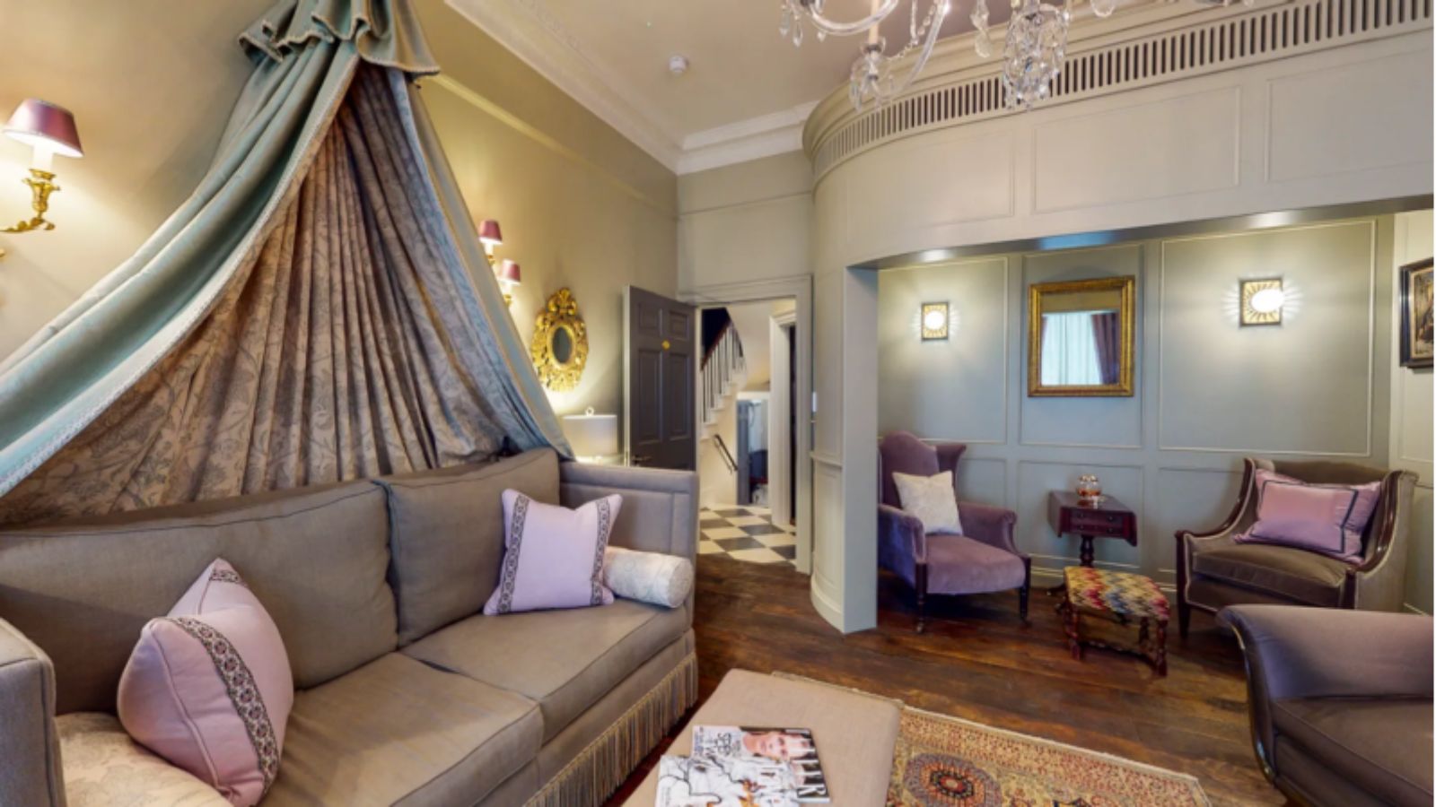 Elegant living room with a gray sofa, draped canopy, purple chairs, ottoman, chandelier, and decorative mirrors on paneled walls; wooden floor and checkered tile entryway visible.