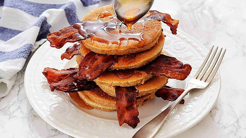 A stack of pancakes with bacon strips between layers is topped with syrup being poured from a spoon, on a white plate with a fork on the side.