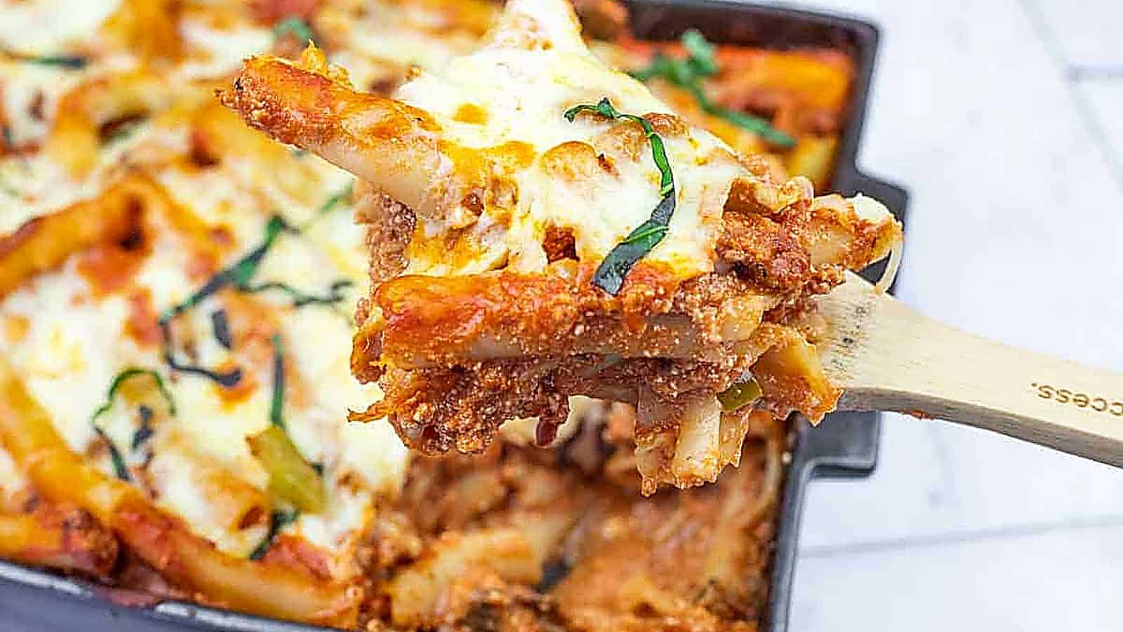 A close-up of a wooden spoon lifting a serving of baked ziti with melted cheese and tomato sauce from a casserole dish.