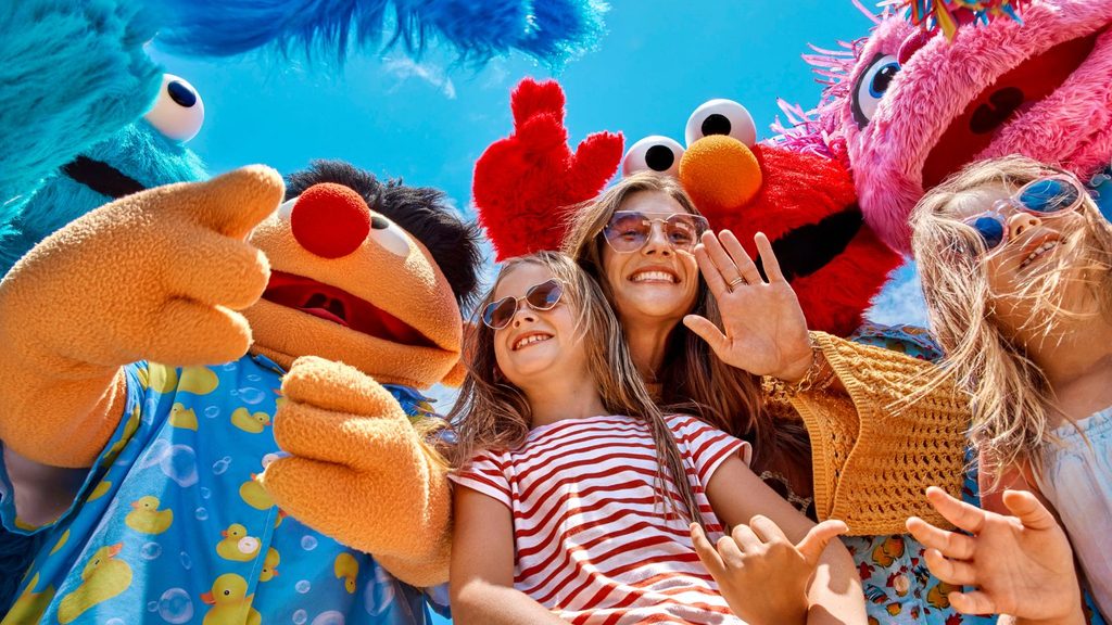 A woman and two children smile and pose with people in colorful Sesame Street character costumes under a bright blue sky.