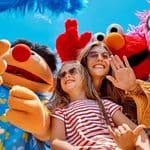 A woman and two children smile and pose with people in colorful Sesame Street character costumes under a bright blue sky.