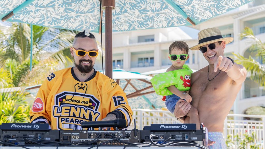 Two men and a child stand by DJ equipment outdoors; one man wears a hockey jersey, the other is shirtless holding the child. All three wear sunglasses under a large umbrella.