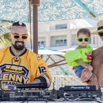 Two men and a child stand by DJ equipment outdoors; one man wears a hockey jersey, the other is shirtless holding the child. All three wear sunglasses under a large umbrella.