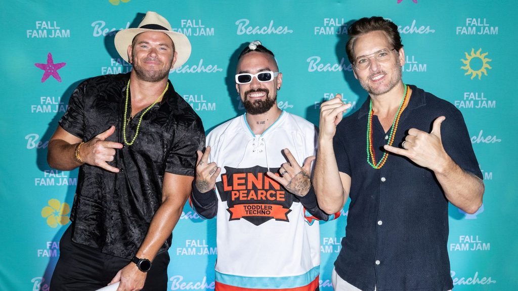 Three men pose in front of a blue “Fall Fam Jam” step-and-repeat banner, wearing casual clothes, sunglasses, hats, and bead necklaces, each making hand gestures and smiling.