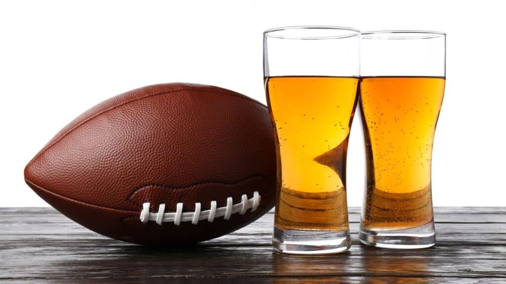 A football rests on a wooden surface next to two tall glasses filled with amber-colored beer, capturing the spirit of NFL stadium beer culture against a white background.