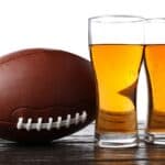 A football rests on a wooden surface next to two tall glasses filled with amber-colored beer, capturing the spirit of NFL stadium beer culture against a white background.