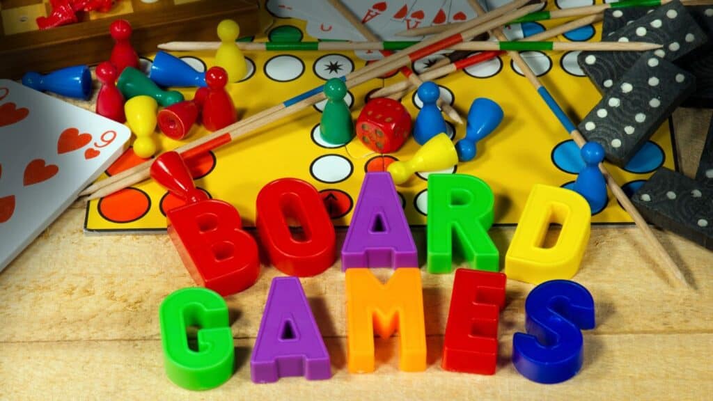 Colorful plastic letters spell “BOARD GAMES” in front of game pieces, cards, dice, and a Ludo board scattered on a wooden surface.