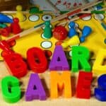 Colorful plastic letters spell &ldquo;BOARD GAMES&rdquo; in front of game pieces, cards, dice, and a Ludo board scattered on a wooden surface.