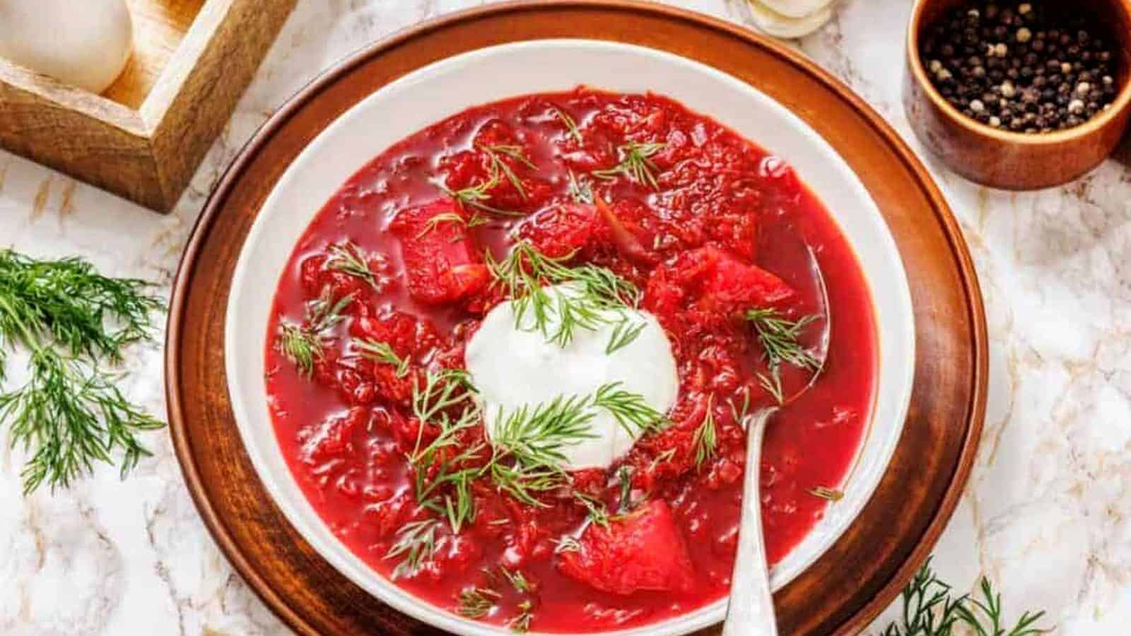 A bowl of borscht with a dollop of sour cream and fresh dill on top, served on a brown plate with a spoon on a marble surface.