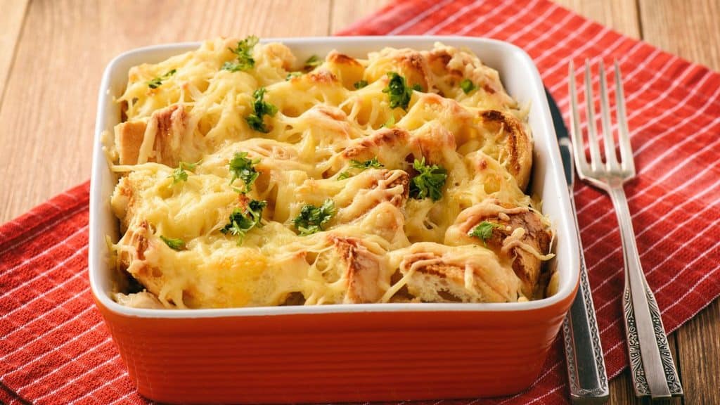 A baked casserole topped with melted cheese and fresh herbs sits in an orange dish on a red striped cloth next to two forks.