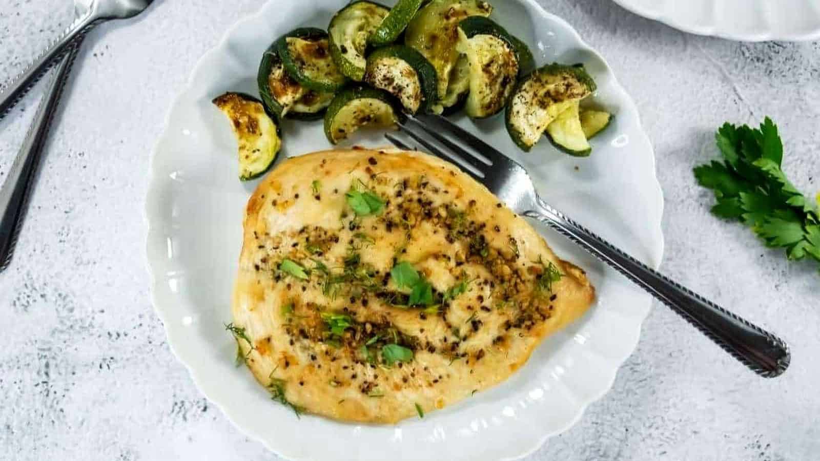A plate with a broiled chicken breast garnished with herbs and a side of roasted zucchini slices, with a fork and knife on the plate.