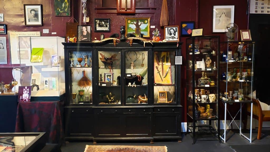 A museum display features a black cabinet and glass case filled with artifacts, artworks, photographs, and curios against red walls, with framed pictures and documents hung above and around them.
