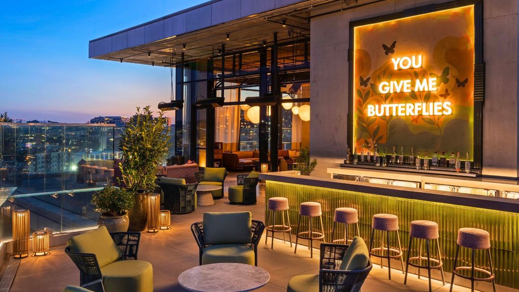 Rooftop bar at sunset with modern seating, greenery, a glass railing, and a neon sign reading “YOU GIVE ME BUTTERFLIES” above the bar.
