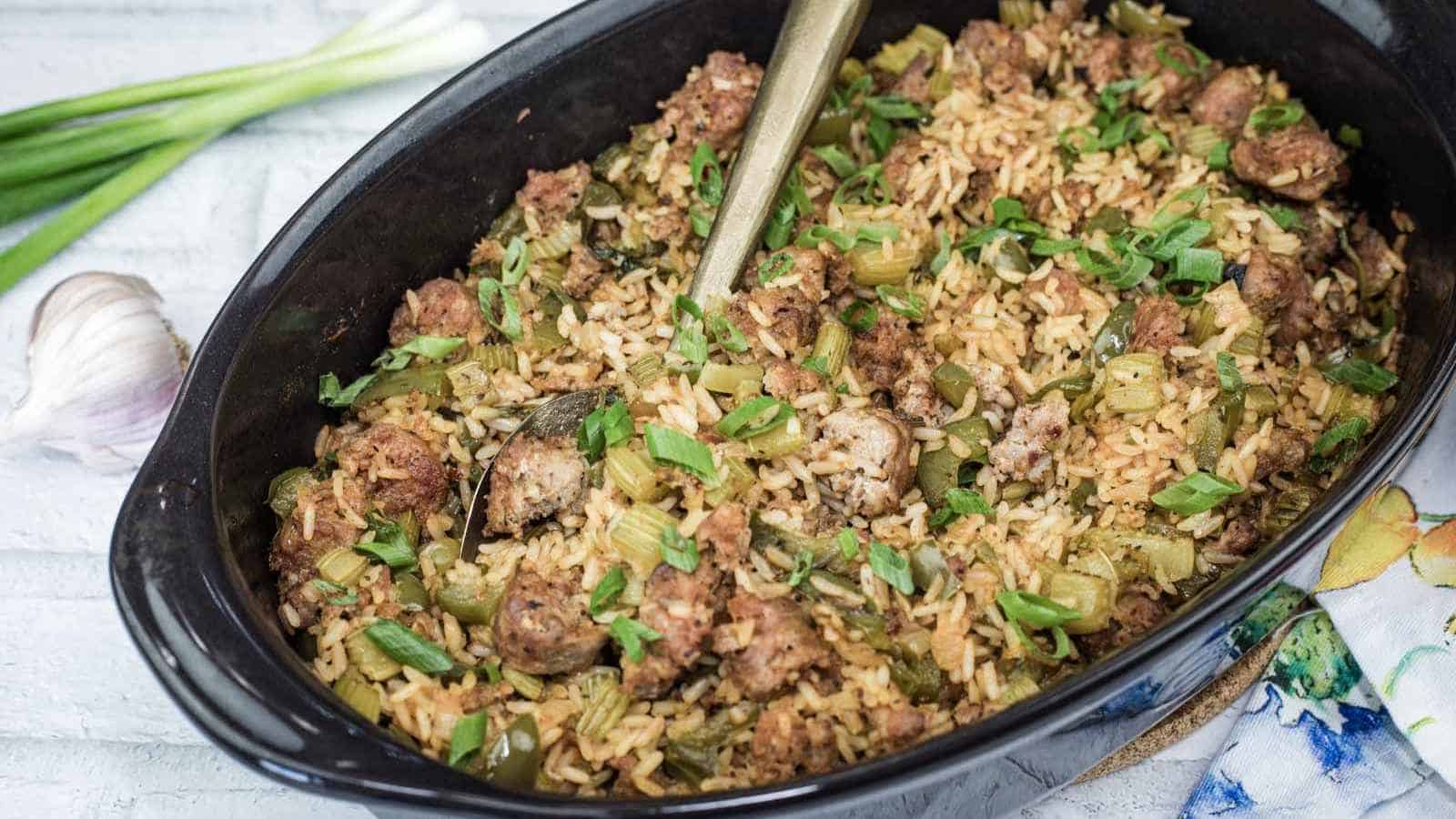 A black dish filled with Cajun Sausage and rice, with a serving spoon inside. Fresh green onions and garlic sit nearby on a white wooden surface.