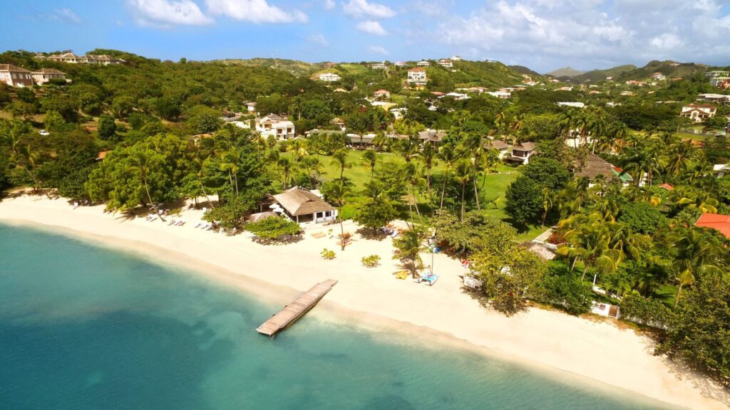 Aerial view of a tropical beach with white sand, calm blue water, a wooden pier, and lush greenery with houses in the background.