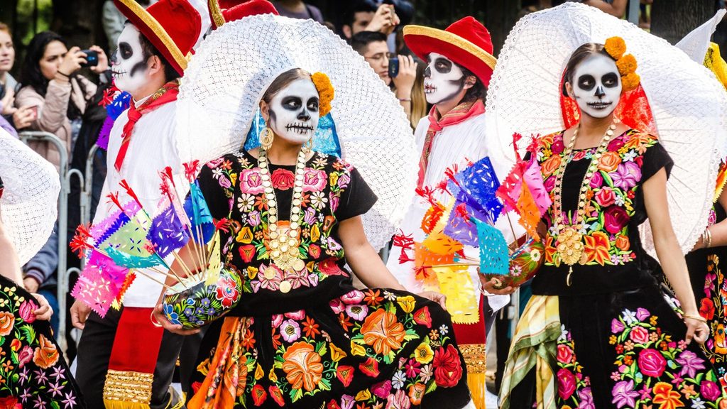People in traditional Mexican clothing with painted skull faces participate in a Day of the Dead parade, holding colorful decorations and wearing large white hats.