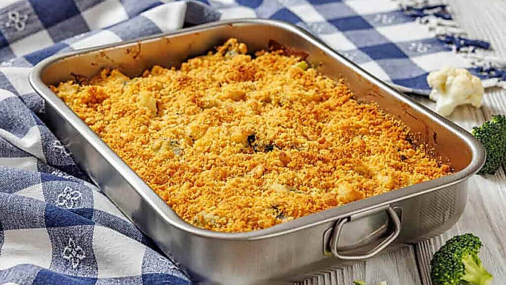 A rectangular metal baking dish filled with a golden brown crumb-topped casserole sits on a blue-and-white checkered cloth. Broccoli and cauliflower are visible around the dish.