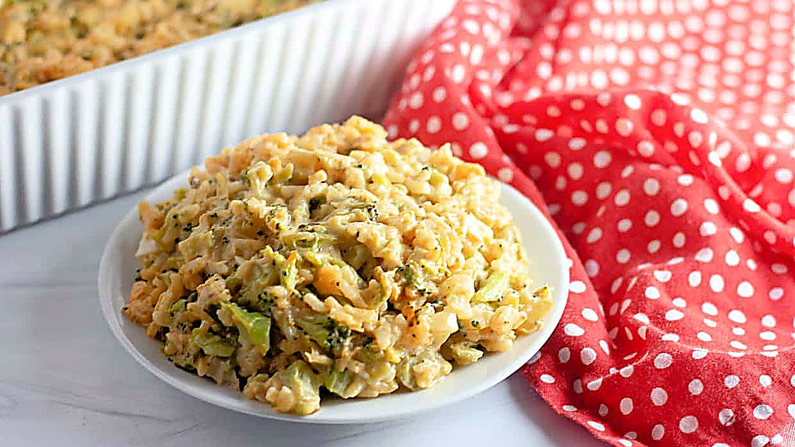 A plate of Easy Cheesy Broccoli Rice Casserole sits on a white surface next to a red and white polka dot cloth, with a baking dish in the background.