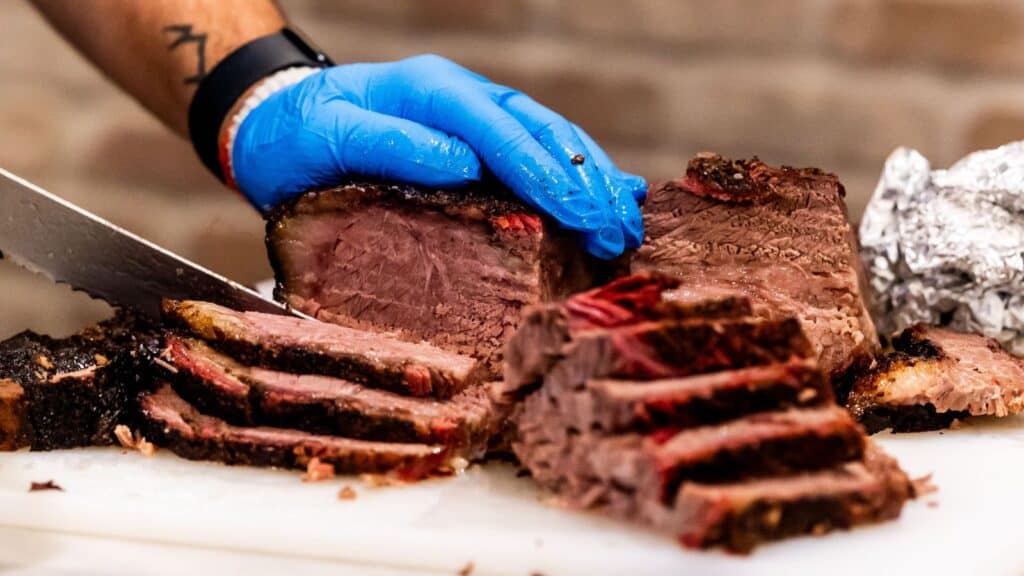 A person wearing a blue glove slices cooked brisket on a white cutting board, with several pieces of meat arranged in front—perfectly capturing the spirit of a Longhorn tailgates brisket contest.