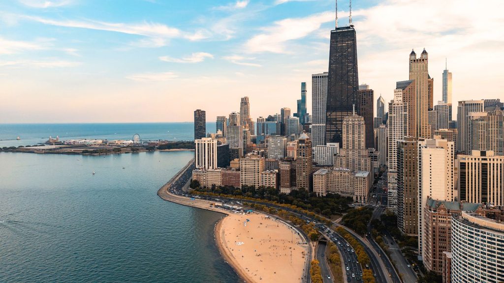 Aerial view of downtown Chicago with skyscrapers, sandy Lake Michigan beach, and a curving lakeside road under a partly cloudy sky.