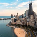 Aerial view of downtown Chicago with skyscrapers, sandy Lake Michigan beach, and a curving lakeside road under a partly cloudy sky.