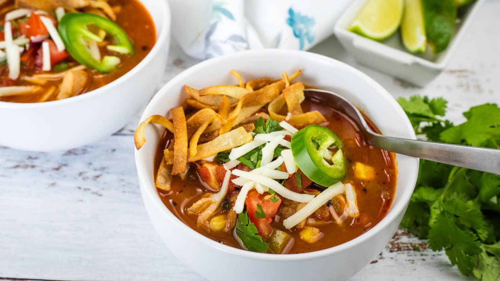 A bowl of chicken tortilla soup topped with shredded cheese, jalape&ntilde;o slices, tortilla strips, and cilantro, with a spoon inside. Another bowl and lime wedges are in the background.