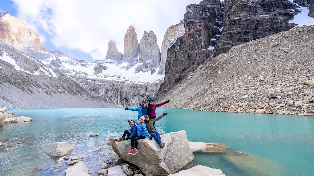Four people pose on rocks beside a turquoise lake with mountains and snowy peaks in the background under a partly cloudy sky.