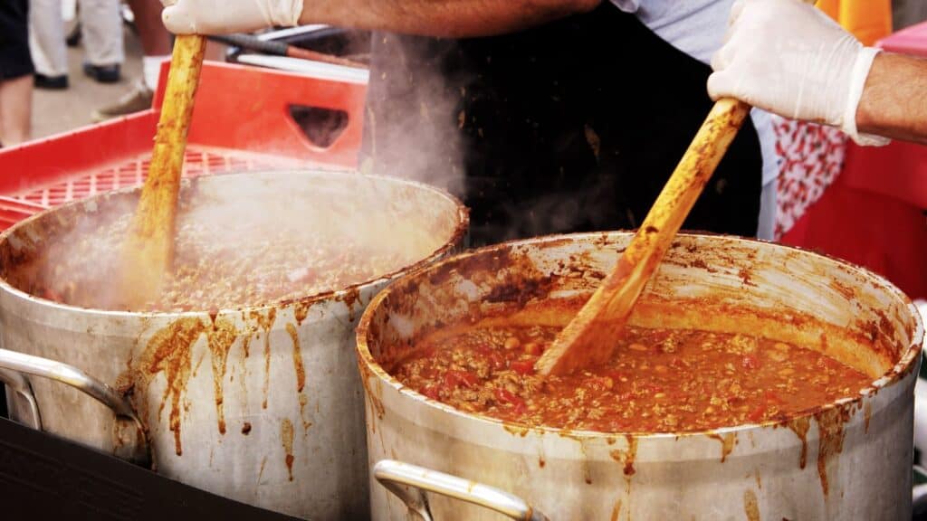 Two large metal pots filled with stew are being stirred by gloved hands using wooden paddles, reminiscent of the hearty preparations seen at fall chili cook-offs. Steam rises from the pots, indicating the food is hot.