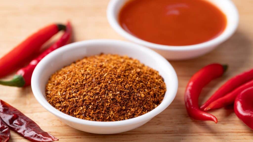 A white bowl of ground chili powder is next to fresh red chili peppers, dried chili pods, and a bowl of red sauce on a wooden surface.