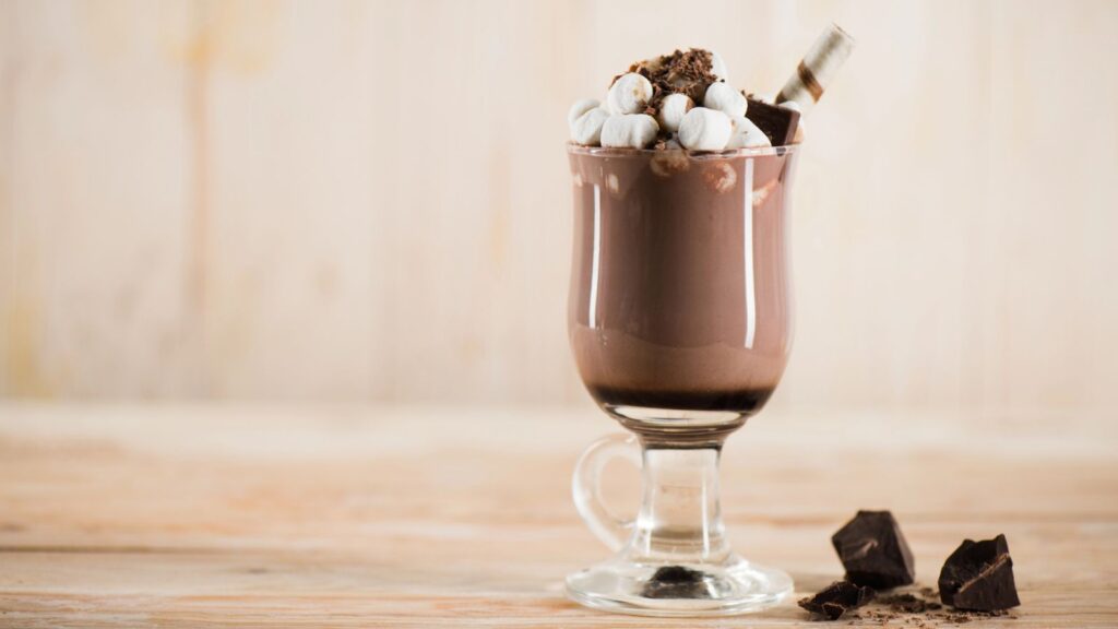 A glass mug of chocolate milkshake topped with mini marshmallows, whipped cream, chocolate shavings, and a chocolate stick sits on a wooden surface with chocolate pieces nearby.