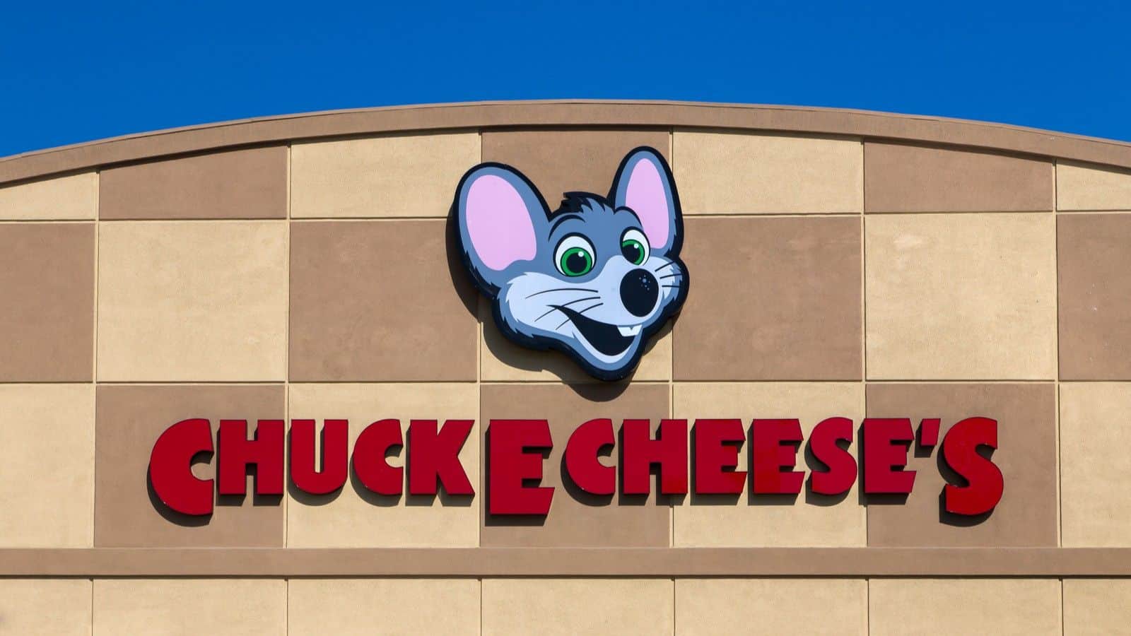 The front of a Chuck E. Cheese’s building with a large cartoon mouse face logo above red lettering on a tan wall against a clear blue sky, representing one of the beloved restaurant chains that came back to delight families once again.