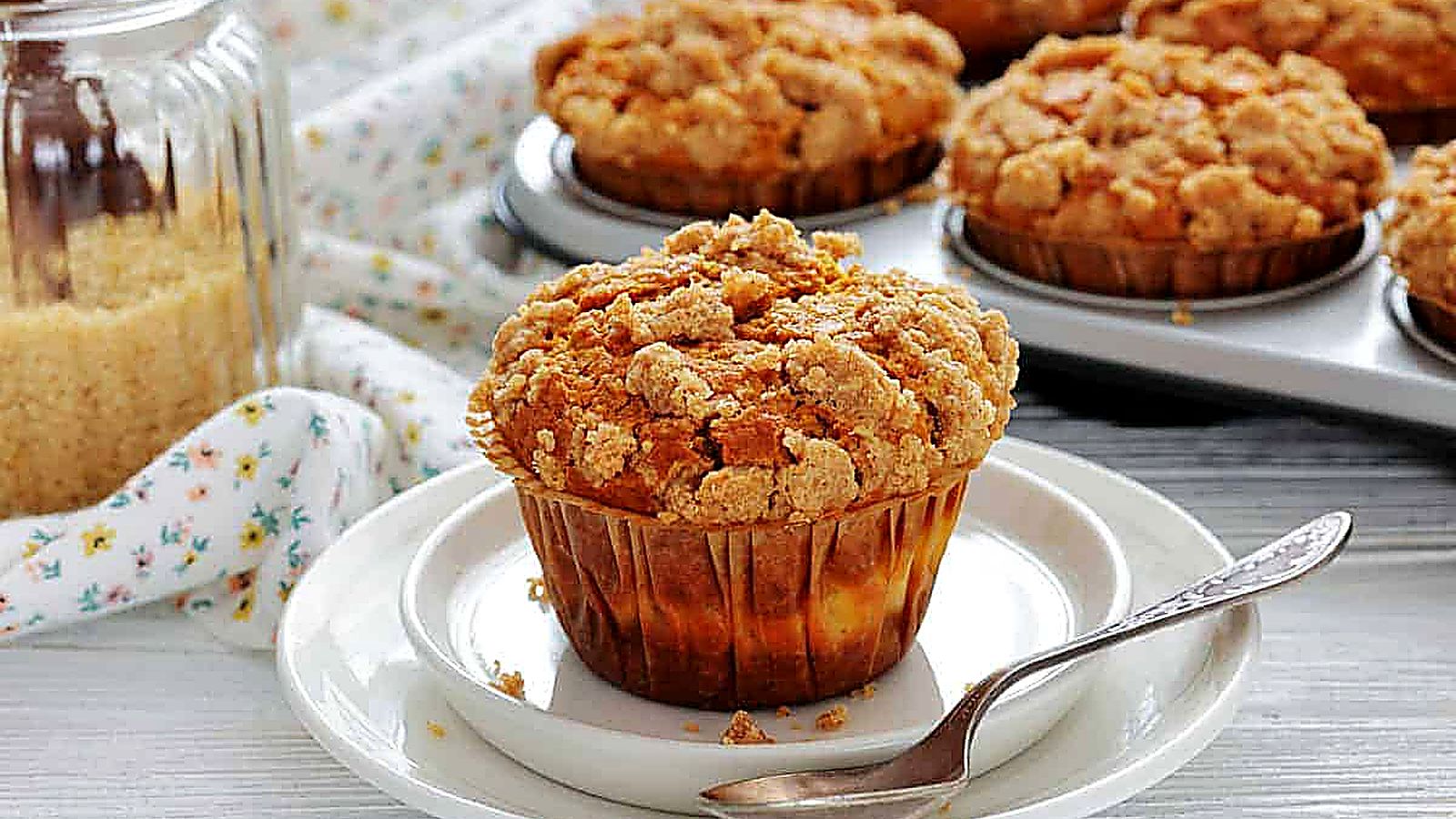 A crumb-topped muffin sits on a white plate with a spoon; more muffins are in a baking tray in the background, next to a jar of brown sugar.
