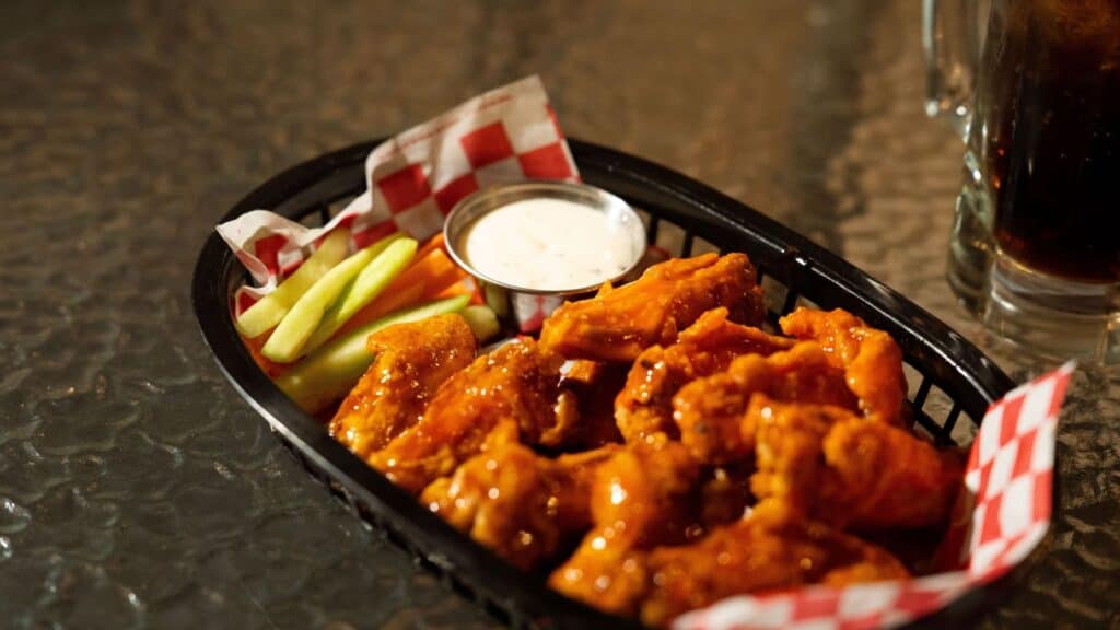 A basket of buffalo chicken wings with ranch dip, celery, and carrot sticks on a checkered paper liner sits on a glass table next to a beverage, inviting you to dig in while you share fun Buffalo wings facts with friends.