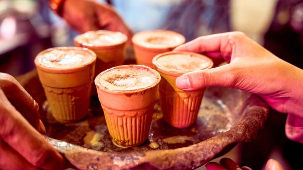 Five clay cups filled with a frothy beverage are placed on a tray, with one hand reaching to pick up a cup&mdash;a perfect scene to celebrate National Chai Day.