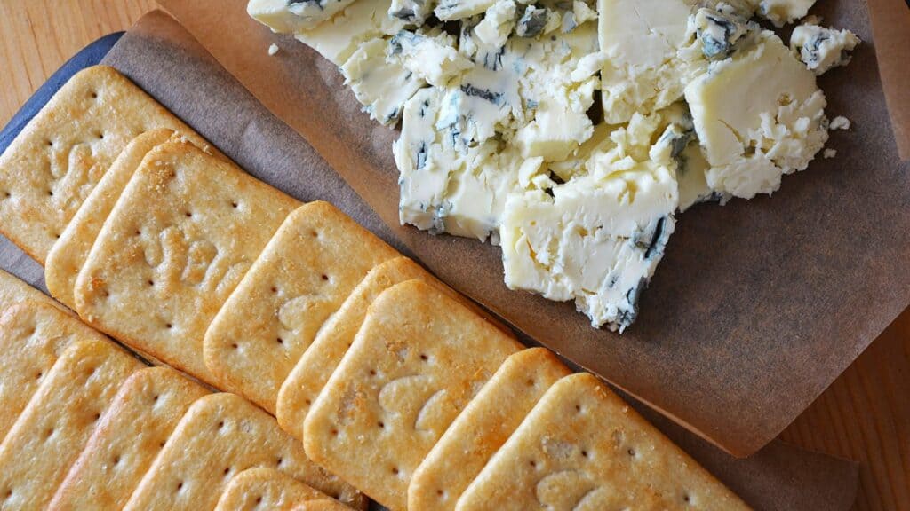 Rectangular crackers arranged next to a pile of crumbled blue cheese on parchment paper atop a wooden surface&mdash;an ideal choice for late-night study snacks.