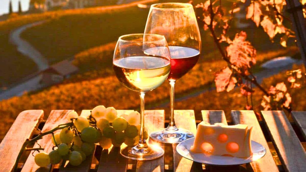 Two glasses of wine, one white and one red, sit on a wooden table with a bunch of grapes and a plate of Swiss cheese, overlooking a sunlit landscape.