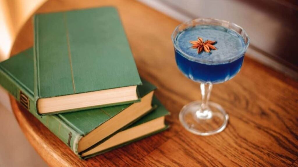 Three green hardcover books&mdash;reminiscent of the volumes an encyclopedia salesman might have offered&mdash;are stacked on a wooden table next to a coupe glass filled with a blue drink, garnished with a star anise.
