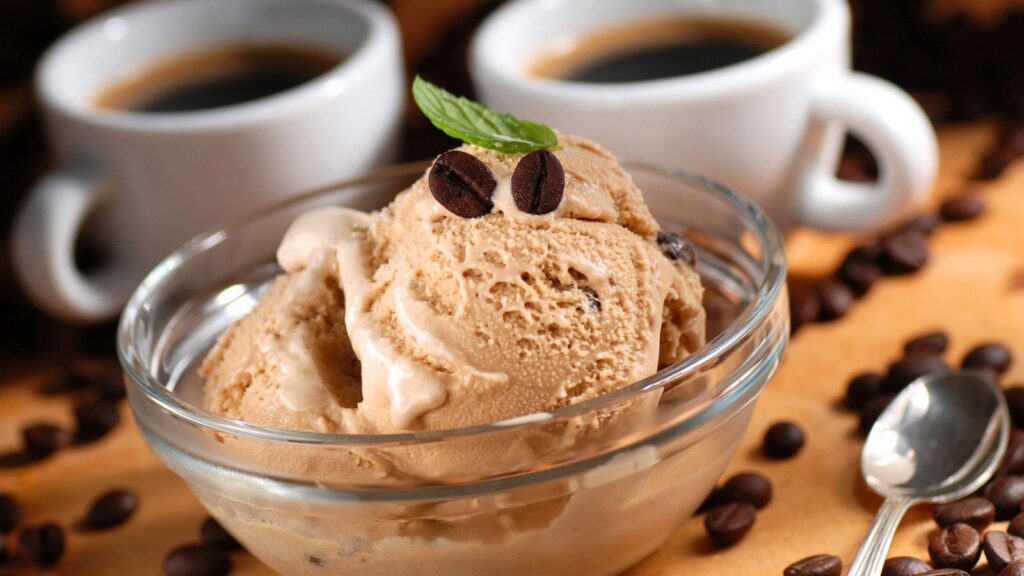A glass bowl of coffee ice cream with coffee beans and a mint leaf on top, next to two cups of coffee, a spoon, and scattered coffee beans.