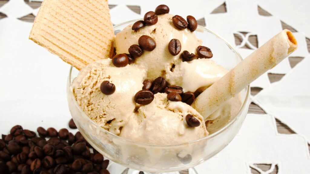 Glass bowl with three scoops of coffee ice cream, topped with coffee beans, a wafer cookie, and a rolled wafer, on a white tablecloth with coffee beans scattered beside the bowl.