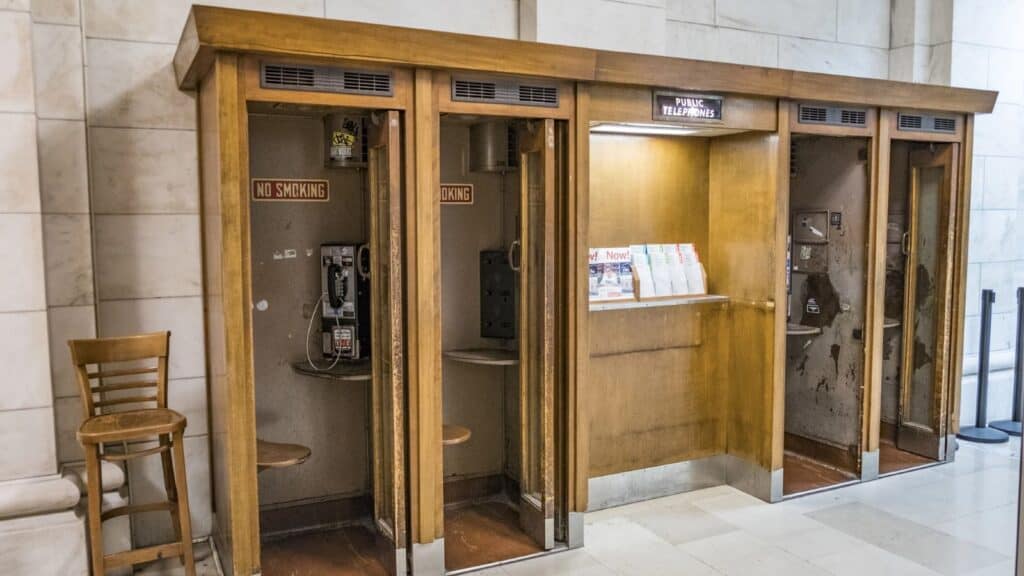 Four vintage wooden phone booths—showcasing payphone booth designs America left behind—stand against a marble wall, three with payphones and one with pamphlets; a wooden chair is placed beside the booths.