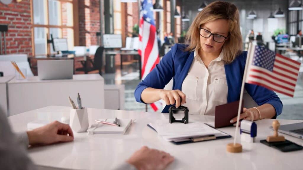 A woman at a desk stamps a document while holding a passport, with an American flag on the table and another flag in the background, emphasizing that US tourists need approval first before proceeding.