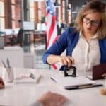 A woman at a desk stamps a document while holding a passport, with an American flag on the table and another flag in the background, emphasizing that US tourists need approval first before proceeding.