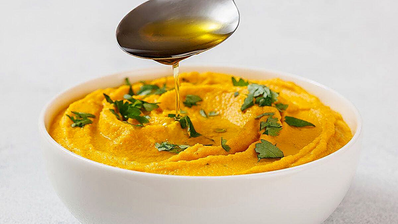 Bowl of Roasted Carrot Hummus garnished with green herbs, with a spoon drizzling oil above it.