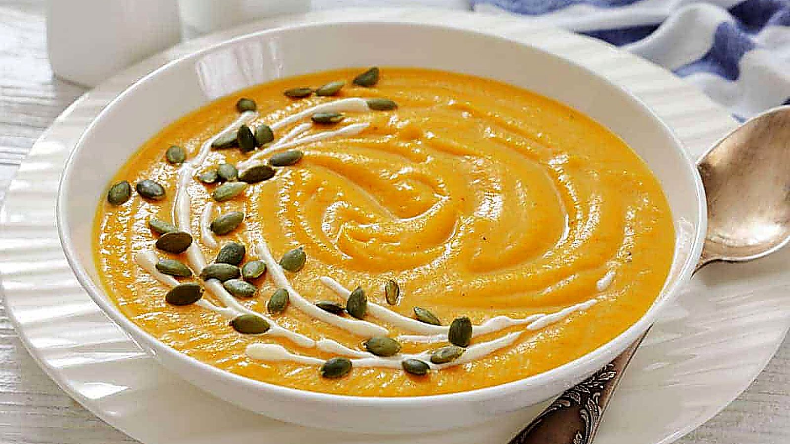 A bowl of creamy pumpkin soup with canned pumpkin garnished with a swirl of cream and sprinkled with pumpkin seeds, placed on a white plate beside a spoon.