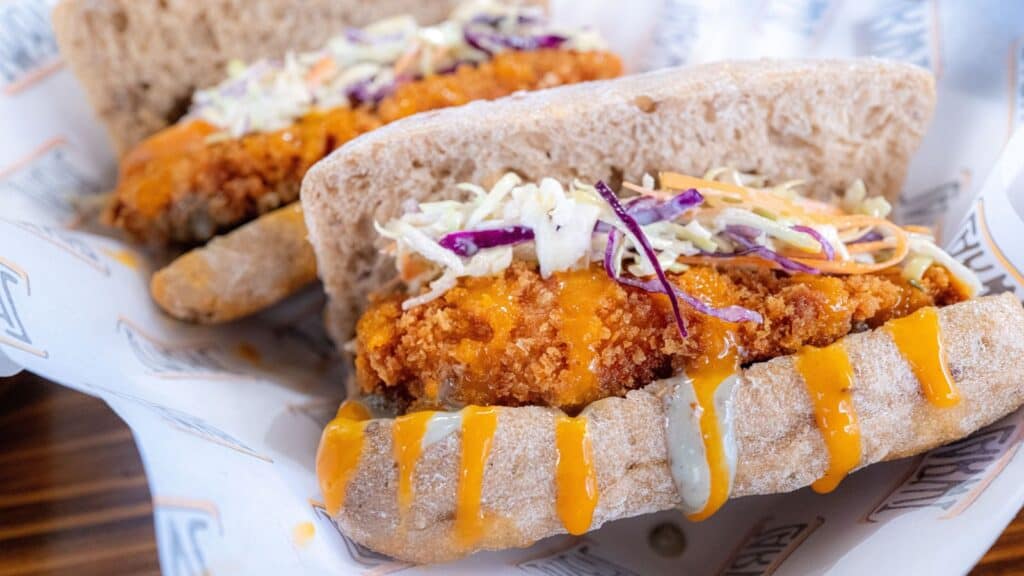 Two breaded chicken sandwiches in buns topped with coleslaw and drizzled with orange sauce, reminiscent of discontinued NFL stadium foods, served on branded paper.
