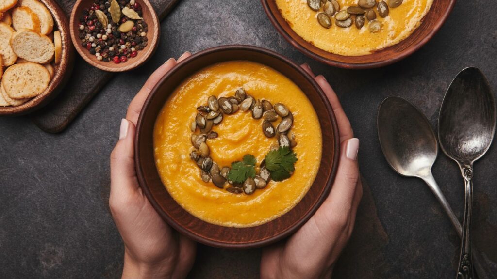 Bowl of creamy orange soup topped with seeds and herbs, held by hands; nearby are a second bowl, two spoons, peppercorns, and toasted bread slices—perfect comfort for soup and stew season.