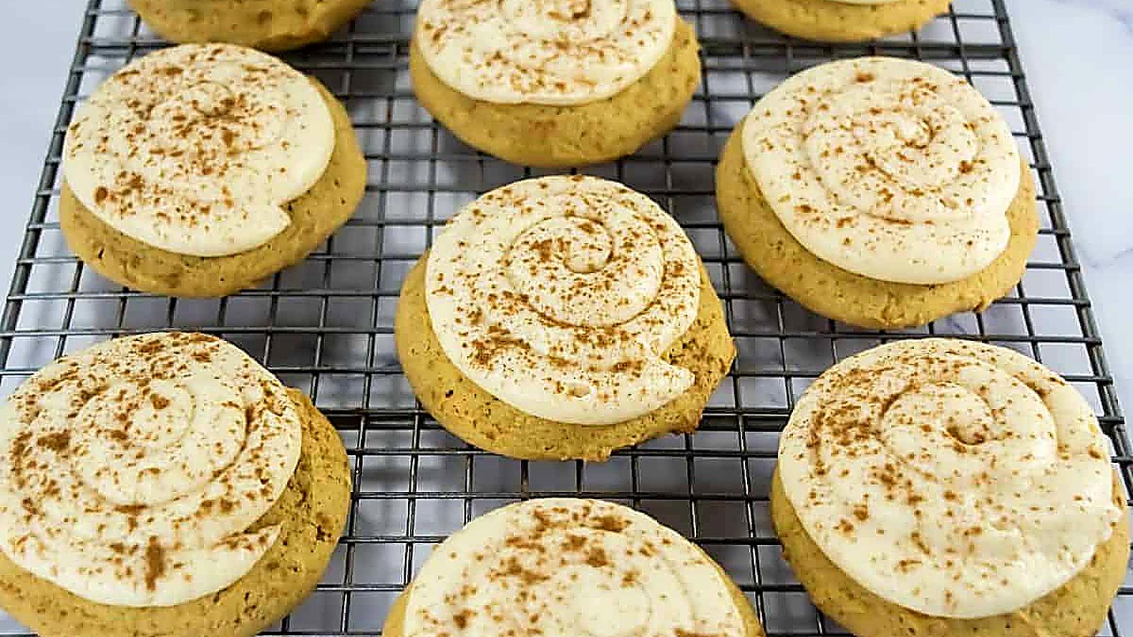 Copycat Crumbl Caramel Pumpkin Cookies with swirled white frosting and a sprinkle of cinnamon are cooling on a wire rack.